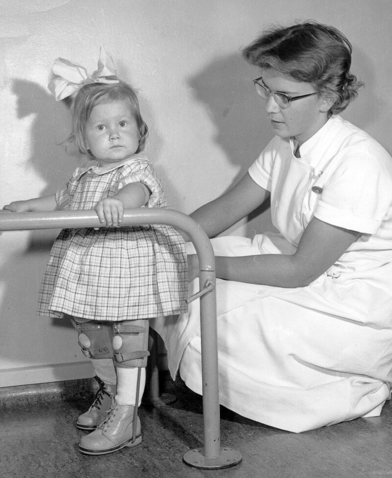 Kuvassa arviolta 3-vuotias tyttö harjoittelee kävelemistä hoitajan ja tukikaiteen avustuksella. Tytön jalka on tuettu, ja hänellä on päässään rusetti.